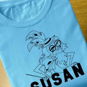May include: A light blue t-shirt with a black and white illustration of a character with a feather boa, hat, and glasses holding a small animal. The text "SUSAN" is printed in black below the illustration.