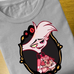 Op de afbeelding: Een grijs T-shirt met een cartoon illustratie. Het ontwerp bevat een personage met een wit gezicht, rode accenten en een zwart kader. Een kleiner varkenspersonage is ook aanwezig, wat bijdraagt aan het algehele ontwerp.