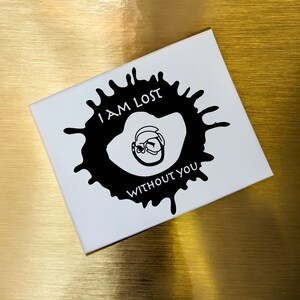 May include: A white card with a black inkblot design. The inkblot has a face inside and the text "I AM LOST WITHOUT YOU".