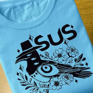May include: Light blue t-shirt with a black graphic design. The design features the word "SUS" above a crow wearing a hat, surrounded by flowers and leaves. The crow has a large eye and is decorated with stars.