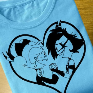May include: A black and white graphic print of two cartoon characters in a heart shape on a light blue t-shirt. The characters are holding hands and looking at each other.