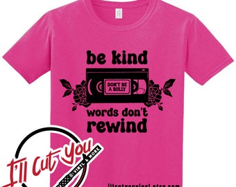 Könnte beinhalten: Pinkes T-Shirt mit einer schwarzen Grafik einer VHS-Kassette mit dem Text "be kind" und "words don't rewind". Das Band hat den Text "Don't be a bully" darauf. Die Grafik ist von schwarzen Blumen umgeben.