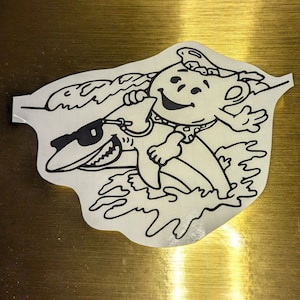 Puede incluir: Una pegatina blanca con un contorno negro que representa a un personaje de dibujos animados surfeando sobre un tiburón. El personaje sonríe y lleva un collar de lunares y un bañador. El tiburón lleva gafas de sol. El diseño tiene un tema de surf.