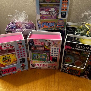 May include: Five colorful toy vending machines with different themes. The machines are decorated with cartoon characters, candy, and other fun designs. Each machine has a button that says "PUSH" and a clear window to see the prizes inside. The themes include "LOL Surprise!", "Sour Patch Kids", "Igor", "Bluey", and "Rilyn's Vending Machine".