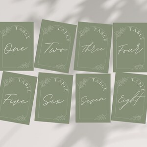 May include: Eight sage green table number cards with white script font. Each card has a decorative arch with the word "TABLE" at the top and a number from one to eight. The cards are decorated with a leaf design.
