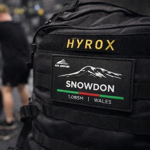 Może przedstawiać: Czarny plecak z żółtym napisem "HYROX". Poniżej naszywka z napisem "SNOWDON" z grafiką gór, wysokością 1085m i "WALES". Zdjęcie zrobione w siłowni.