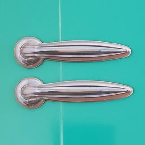 May include: Two silver cabinet door handles with a rounded, slightly curved design. The handles are against a turquoise background.
