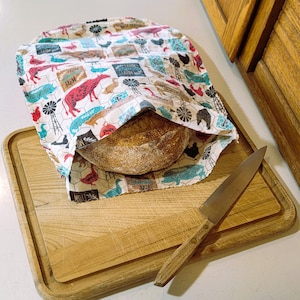 May include: A fabric bread bag with a white background and a multi-colored farm-themed print featuring cows, chickens, windmills, and the word farm. A round loaf of crusty bread is inside the bag, which rests on a wooden cutting board.