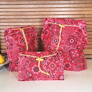May include: Three fabric pouches in varying sizes featuring a red and black paisley bandana print. Each pouch is secured with a beige drawstring cord and a small yellow wooden toggle closure, providing a consistent look across the set.