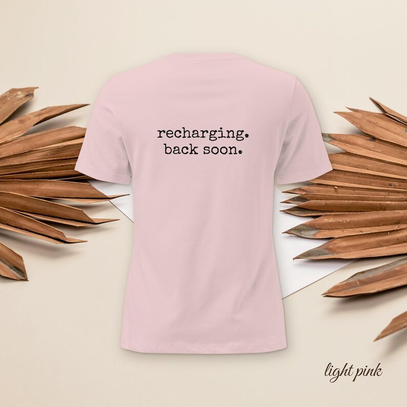 May include: A light pink short-sleeved t-shirt featuring the black text "recharging. back soon." printed in a typewriter-style font on the upper center of the garment. The shirt has a crew neckline and a plain, solid-colored back design.