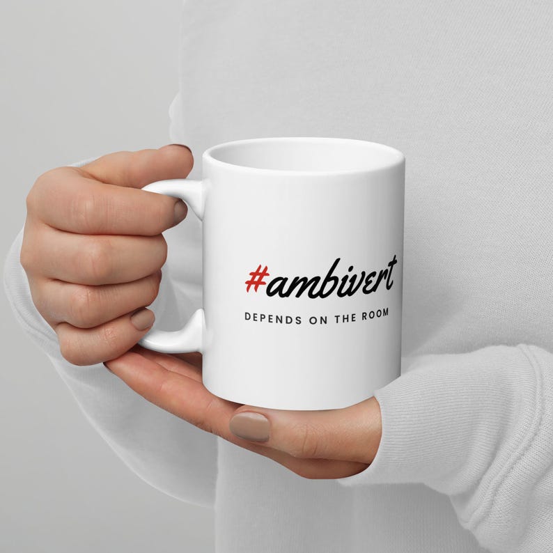 May include: A white ceramic mug featuring the red hashtag symbol followed by the black text #ambivert and the phrase DEPENDS ON THE ROOM printed in black capital letters below it. The mug has a standard handle and a smooth, cylindrical body.