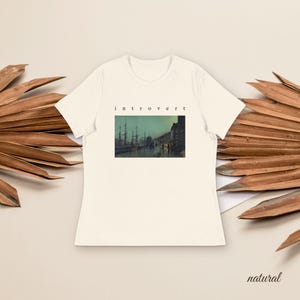May include: A cream-colored short-sleeve t-shirt featuring a rectangular graphic print of a dark harbor scene with ships and buildings. The word "introvert" is printed in small black lowercase letters centered above the rectangular image.