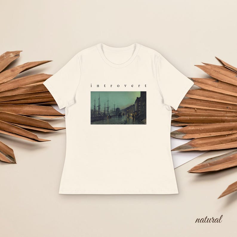 May include: A cream-colored short-sleeve t-shirt featuring a rectangular graphic print of a dark harbor scene with ships and buildings. The word "introvert" is printed in small black lowercase letters centered above the rectangular image.