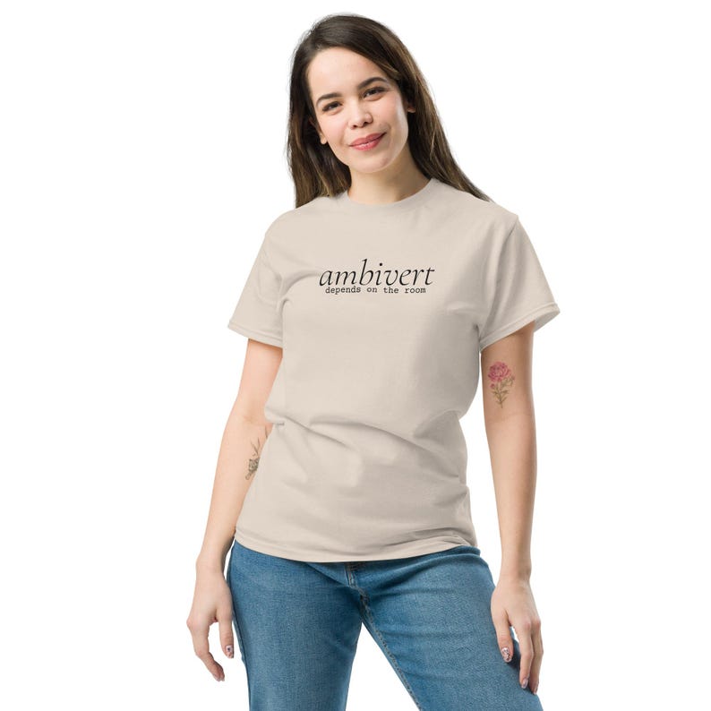 May include: A beige short-sleeved t-shirt featuring the black text "ambivert" in a serif font with the smaller text "depends on the room" underneath. The shirt is paired with blue denim jeans, creating a casual and neutral-toned outfit.