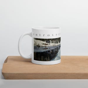 Puede incluir: Una taza de cer&aacute;mica blanca con una impresi&oacute;n de un paisaje de ribera nevada con agua oscura que refleja los &aacute;rboles y el cielo. La palabra introvert est&aacute; impresa en una fuente negra en min&uacute;sculas sobre la imagen del paisaje.