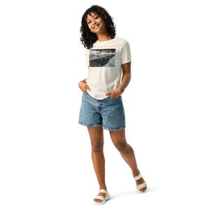 May include: A white short-sleeved t-shirt featuring a rectangular graphic print of a snowy landscape with the text "WILDLIFE" above it, paired with blue denim shorts and white sandals. The t-shirt has a crew neckline and a standard fit.