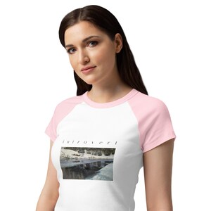 May include: A white raglan t-shirt with light pink sleeves and a crew neckline. The front features a rectangular photographic print of a snowy landscape with a dark body of water and the word "introvert" printed in black lowercase letters above it.