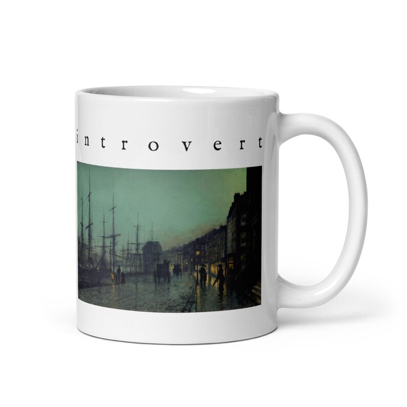 May include: A white ceramic mug featuring the word "introvert" printed in black lowercase letters above a dark, atmospheric painting of a harbor scene at dusk with tall ship masts, silhouettes of buildings, and figures on a wet cobblestone street.