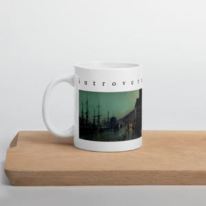 May include: A white ceramic mug featuring the word "introvert" in black lowercase letters above a dark, moody illustration of a harbor at night with silhouetted ships, masts, and buildings reflected in the water under a greenish sky.