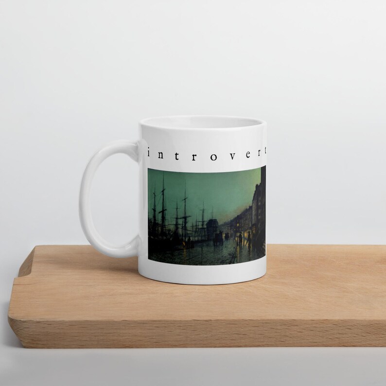 May include: A white ceramic mug featuring the word "introvert" in black lowercase letters above a dark, moody illustration of a harbor at night with silhouetted ships, masts, and buildings reflected in the water under a greenish sky.