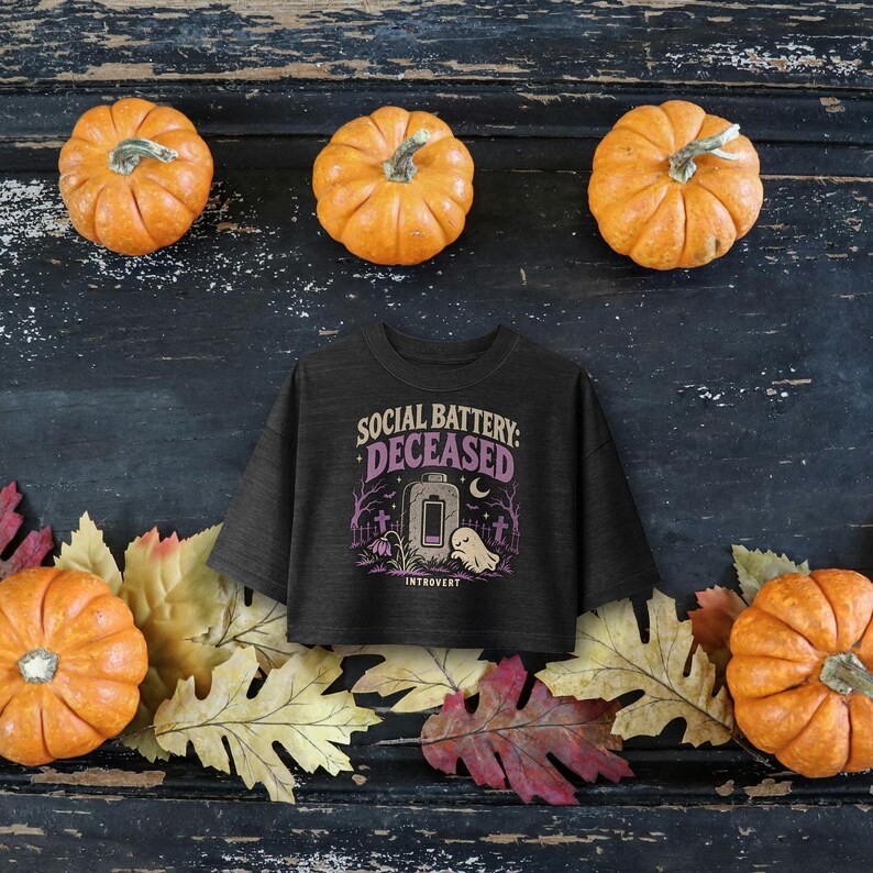 May include: A dark gray cropped t-shirt featuring a graphic print with the text SOCIAL BATTERY: DECEASED, an illustration of a low battery icon, a ghost, tombstones, and trees, all rendered in shades of purple, white, and light gray.