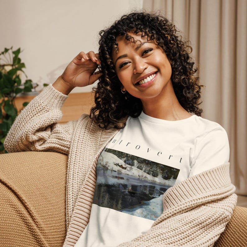 May include: A white t-shirt featuring a rectangular graphic print of a snowy landscape with a dark lake and the word "introvert" printed in black, sans-serif capital letters above the image. The shirt is worn under a beige knit cardigan.