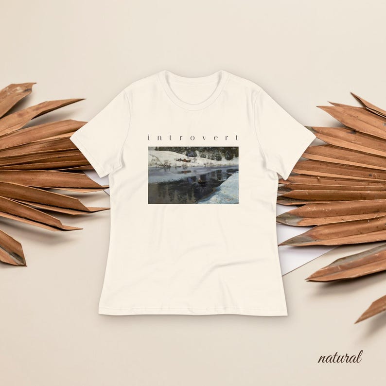 May include: A cream-colored short-sleeve t-shirt featuring a rectangular graphic print of a snowy river landscape. Above the image, the word "introvert" is printed in a simple black sans-serif font. The shirt has a crew neckline and hemmed sleeves.