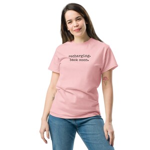 May include: A light pink short-sleeved t-shirt featuring the black text "recharging. back soon." printed in a typewriter-style font across the chest. The garment has a crew neckline and a standard fit, paired with blue denim jeans.