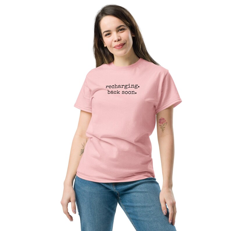 May include: A light pink short-sleeved t-shirt featuring the black text "recharging. back soon." printed in a typewriter-style font across the chest. The garment has a crew neckline and a standard fit, paired with blue denim jeans.
