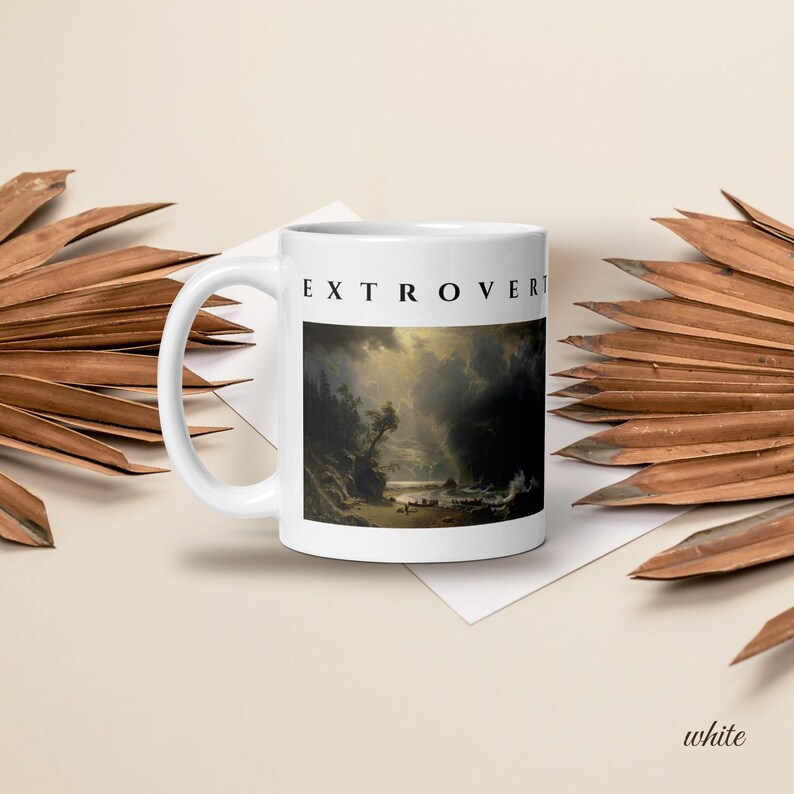 May include: A white ceramic mug featuring the word EXTROVERT printed in black capital letters above a rectangular landscape painting depicting a dark, stormy sky over a coastal scene with trees, cliffs, and a small boat on the water.
