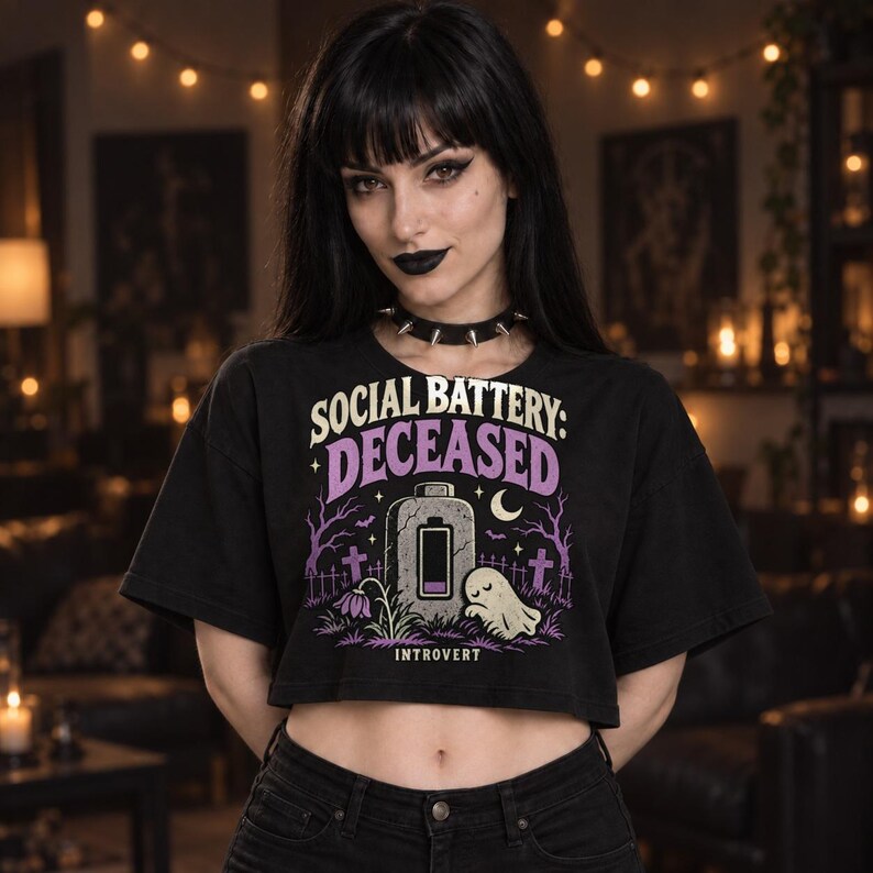 May include: A black cropped t-shirt featuring a graphic print with the text "SOCIAL BATTERY: DECEASED" and "INTROVERT". The design includes a tombstone shaped like a battery, a small ghost, purple trees, and crosses in a graveyard setting.