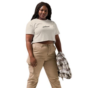 May include: A beige short-sleeved crop top featuring the word ambivert printed in a black cursive script across the chest. The garment is paired with tan cargo pants and a brown and white plaid flannel shirt held in one hand.