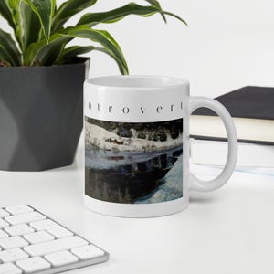 Puede incluir: Una taza de cer&aacute;mica blanca con la palabra introvert impresa en letras min&uacute;sculas negras sobre una ilustraci&oacute;n de un paisaje con una orilla nevada, una peque&ntilde;a caba&ntilde;a roja y un cuerpo de agua oscuro y reflectante.