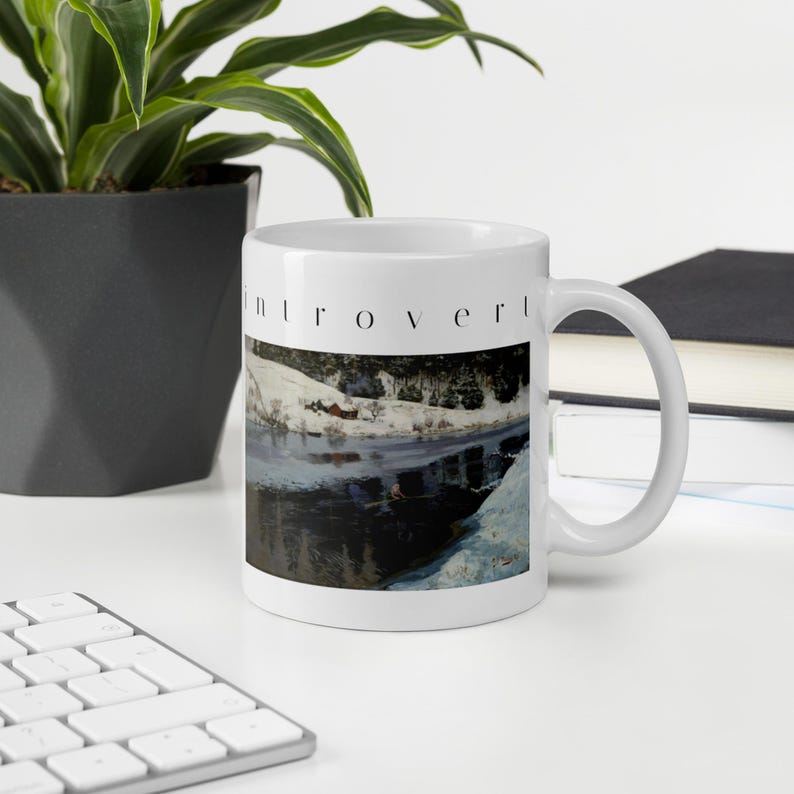 Puede incluir: Una taza de cer&aacute;mica blanca con la palabra introvert impresa en letras min&uacute;sculas negras sobre una ilustraci&oacute;n de un paisaje con una orilla nevada, una peque&ntilde;a caba&ntilde;a roja y un cuerpo de agua oscuro y reflectante.