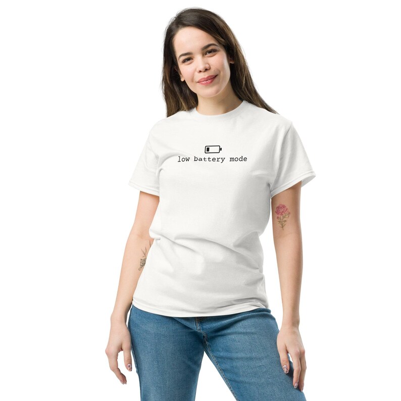 May include: A white short-sleeved t-shirt featuring a black graphic of a battery icon and the text "low battery mode" printed in a typewriter-style font across the center of the chest area, paired with blue denim jeans.