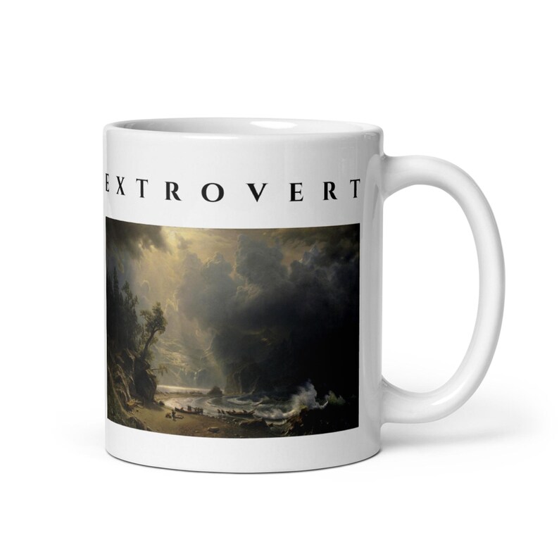 May include: A white ceramic mug featuring the black text EXTROVERT printed above a dark, dramatic landscape painting depicting a stormy coastal scene with crashing waves, rocky cliffs, and light breaking through heavy, dark clouds.