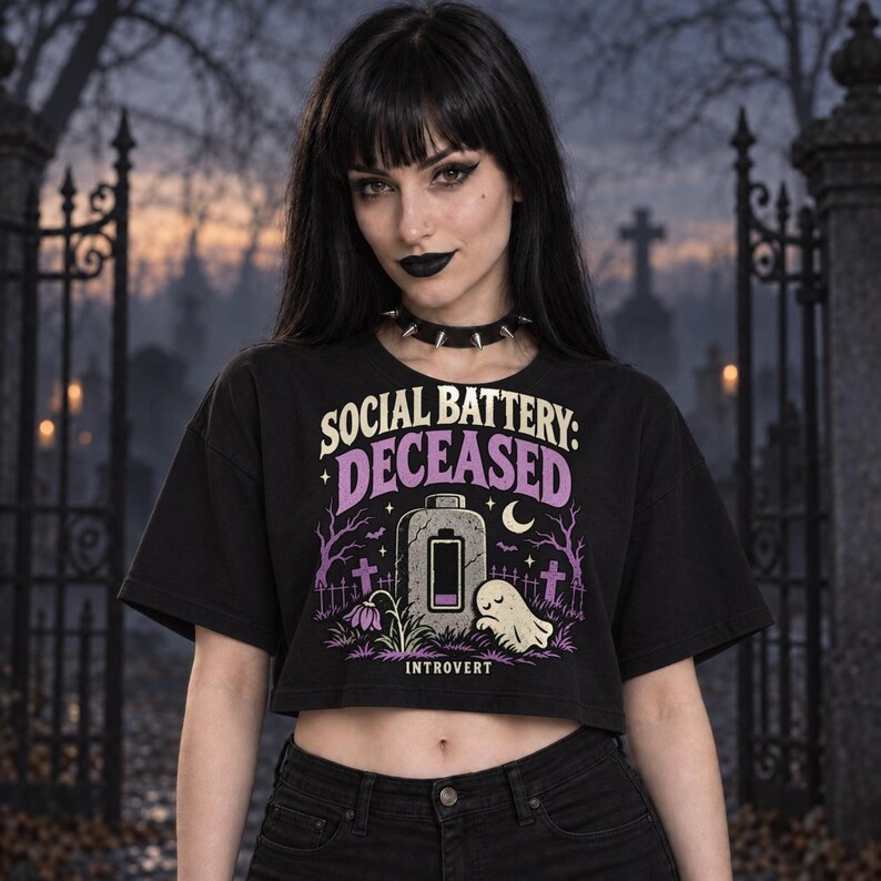 May include: A black cropped t-shirt featuring a graphic print with the text "SOCIAL BATTERY: DECEASED" and "INTROVERT". The design includes a tombstone with an empty battery icon, a small ghost, bare trees, and a crescent moon in purple and cream.