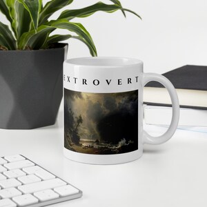 May include: A white ceramic mug featuring the word EXTROVERT printed in black capital letters above a dark, dramatic landscape painting depicting a stormy coastal scene with turbulent clouds, rocky cliffs, and a small boat on the water.