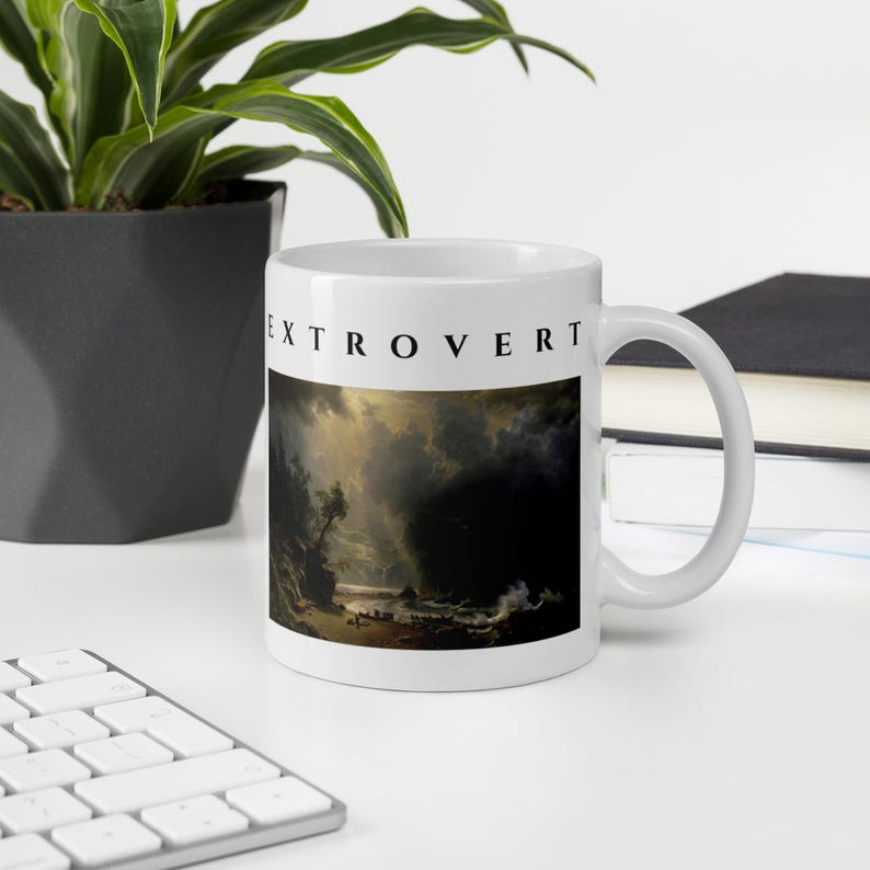 May include: A white ceramic mug featuring the word EXTROVERT printed in black capital letters above a dark, dramatic landscape painting depicting a stormy coastal scene with turbulent clouds, rocky cliffs, and a small boat on the water.