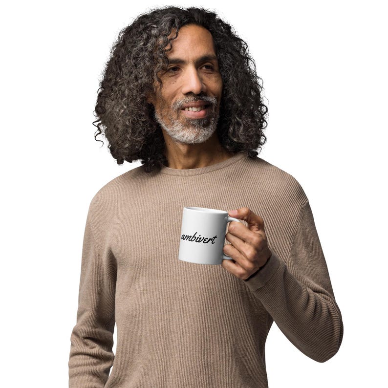 May include: A white ceramic mug featuring the word ambivert printed in a black cursive script font. The mug has a standard cylindrical shape with a curved handle on the side, designed for holding hot or cold beverages in a simple, functional style.
