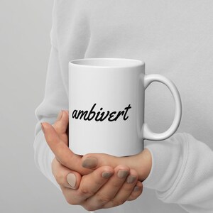 May include: A white ceramic mug featuring the word ambivert printed in a black cursive script font. The mug has a standard cylindrical shape with a curved handle on the right side, providing a clean and simple design for daily beverage consumption.