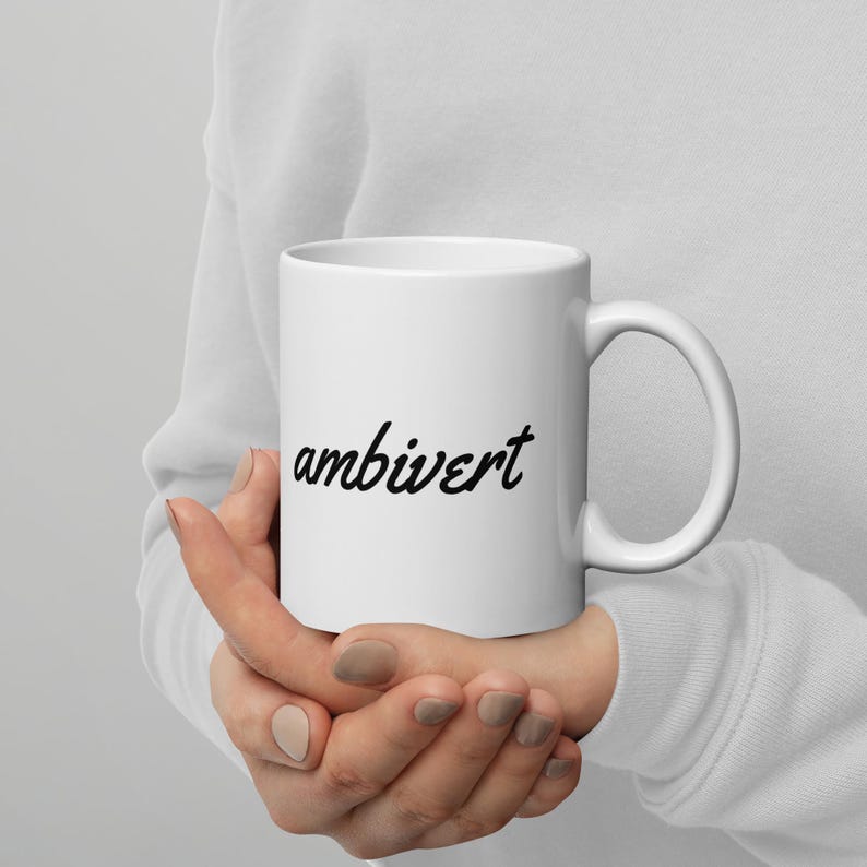 May include: A white ceramic mug featuring the word ambivert printed in a black cursive script font. The mug has a standard cylindrical shape with a curved handle on the right side, providing a clean and simple design for daily beverage consumption.