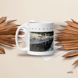 Puede incluir: Una taza de cer&aacute;mica blanca con la palabra introvert impresa en fuente negra sin remates sobre una imagen rectangular de un paisaje con una orilla nevada, un peque&ntilde;o edificio rojo y agua oscura que refleja el entorno invernal.