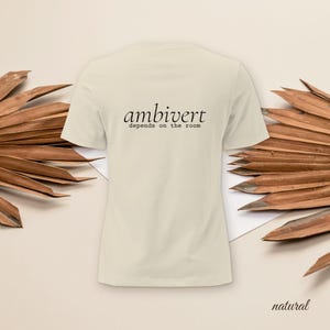 May include: A natural-colored short-sleeve t-shirt featuring the black text "ambivert" in a stylized serif font, with the phrase "depends on the room" printed in a smaller, typewriter-style font directly underneath the main word.