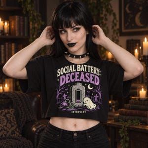 May include: A black cropped t-shirt featuring a graphic print with the text "SOCIAL BATTERY: DECEASED" and "INTROVERT". The design includes a battery icon, a ghost, a crescent moon, and a graveyard scene in shades of purple, white, and grey.