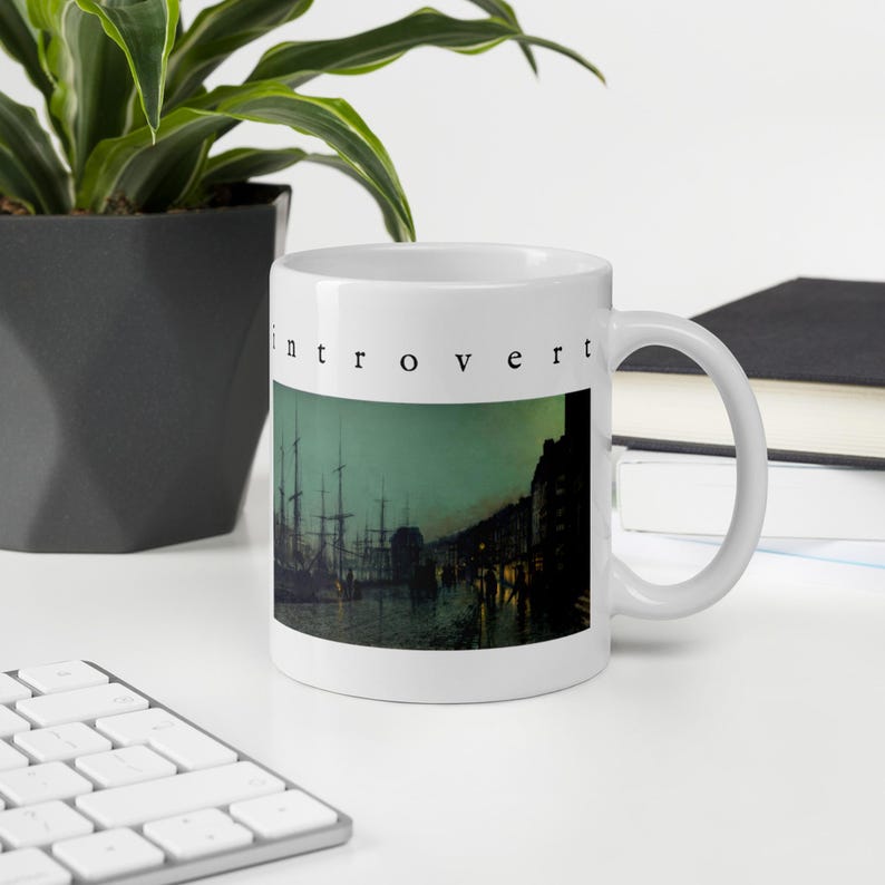 May include: A white ceramic mug featuring a printed image of a dark harbor scene with ships at night. The word "introvert" is printed in a black, lowercase serif font above the artwork. The mug has a standard handle and a smooth finish.