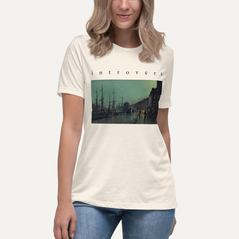 May include: A cream-colored short-sleeve t-shirt featuring the word "introvert" printed in black text above a rectangular graphic depicting a dark, moody harbor scene with tall ship masts, calm water, and dim lighting along the shoreline.