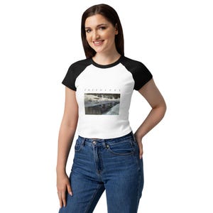 May include: A white raglan t-shirt with black sleeves and a black crew neckline. The center of the shirt features a rectangular graphic print of a snowy landscape with the word "introvert" printed in a small, black, sans-serif font above it.