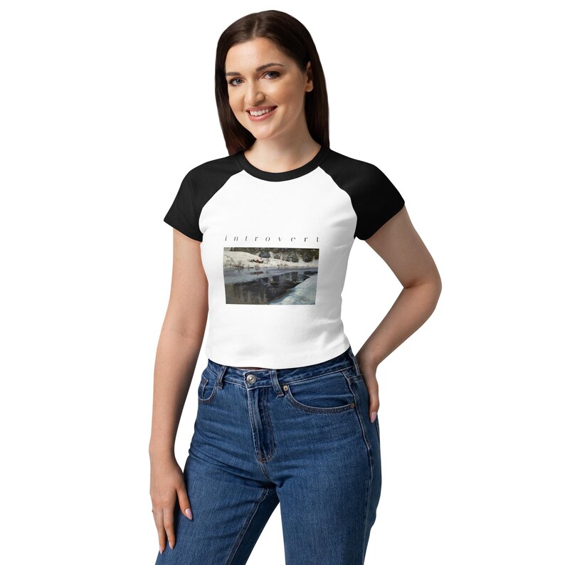 May include: A white raglan t-shirt with black sleeves and a black crew neckline. The center of the shirt features a rectangular graphic print of a snowy landscape with the word "introvert" printed in a small, black, sans-serif font above it.
