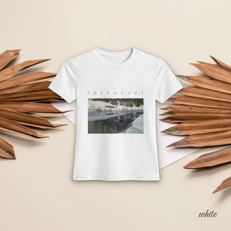 May include: A white short-sleeved t-shirt featuring a rectangular graphic print of a snowy landscape with a dark river and the word "introvert" printed in black, lowercase, spaced-out lettering centered above the image.